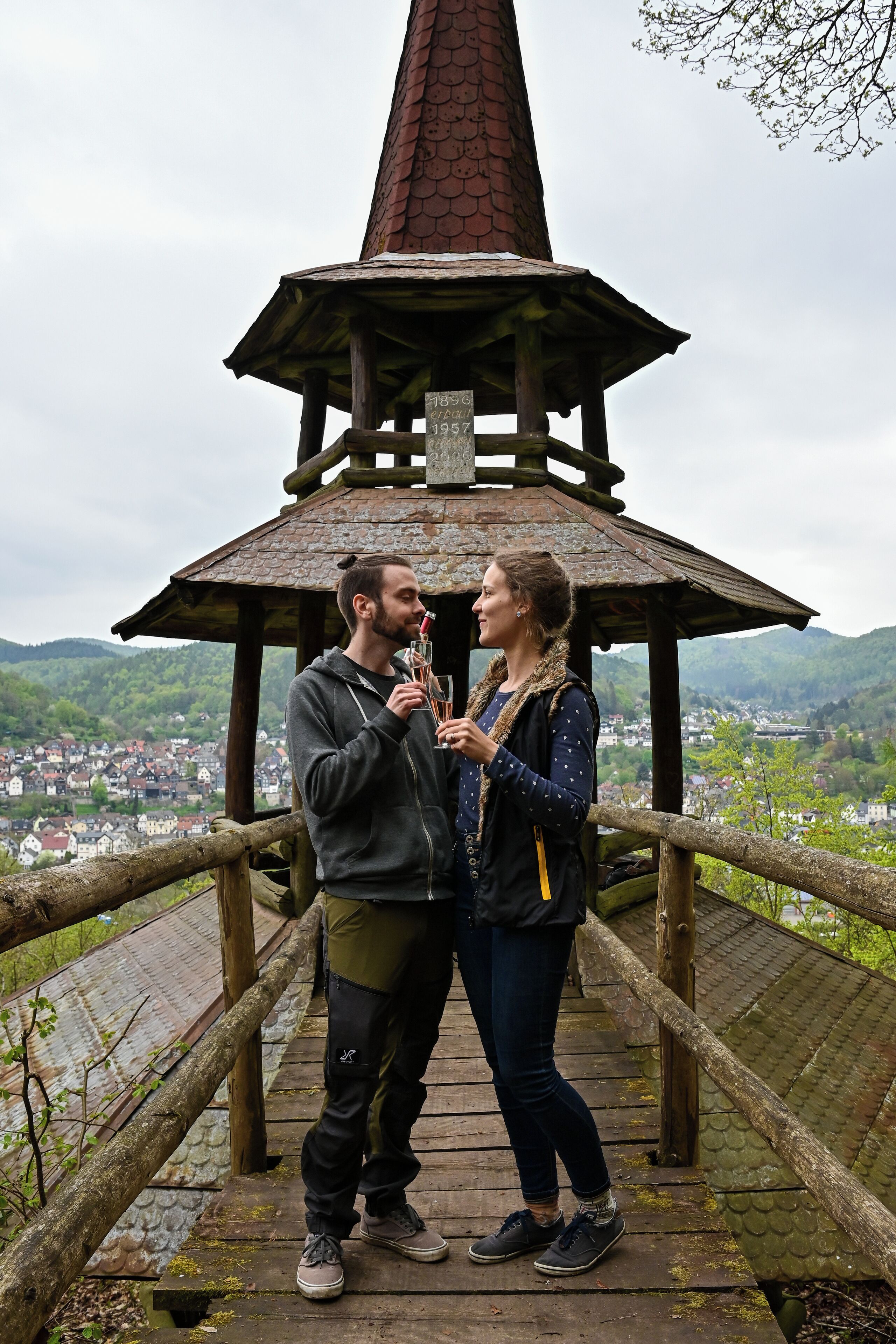 A couple standing on a wooden bridge with a view of a town and hills in the background.