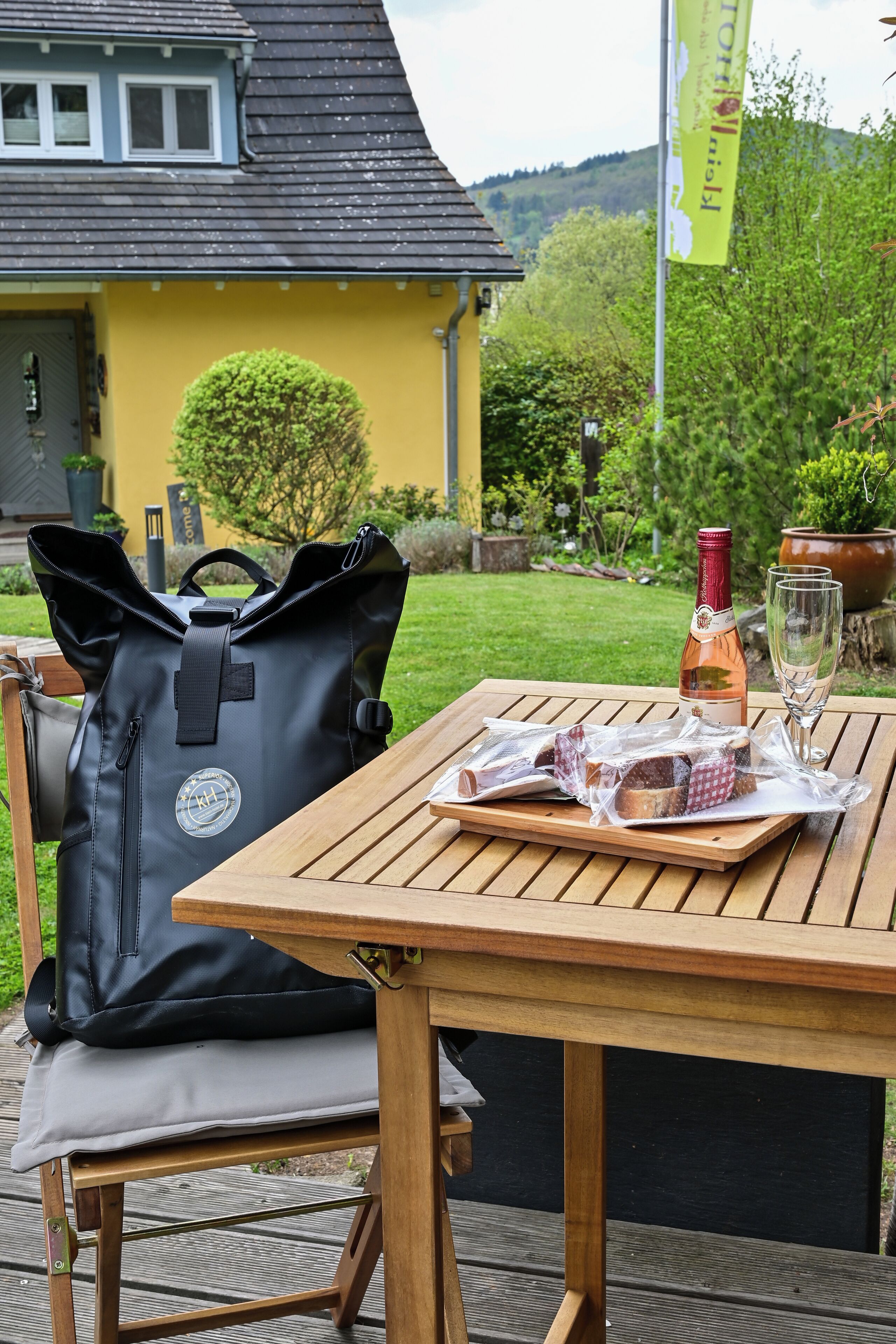 A wooden table with a bottle of wine and glasses, a black bag, and a cushion on a deck.