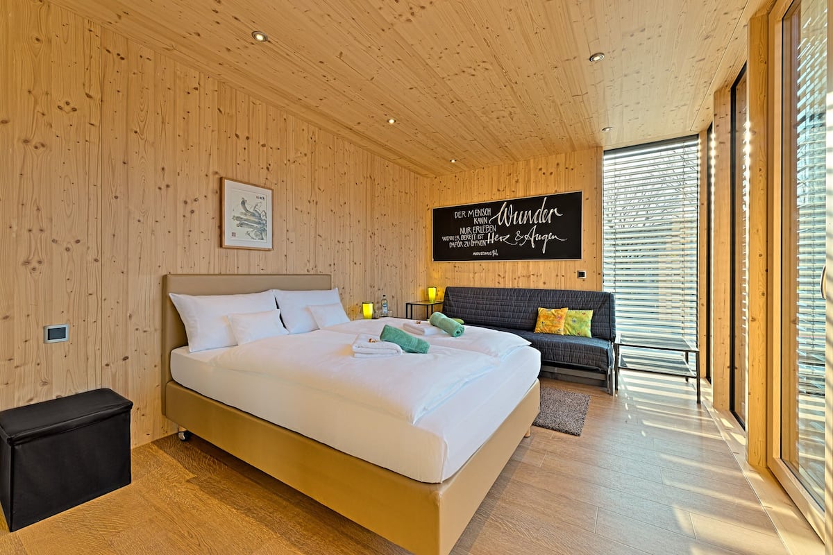 A modern hotel room with a wooden interior, a large bed, a sofa, and a wall-mounted TV.
