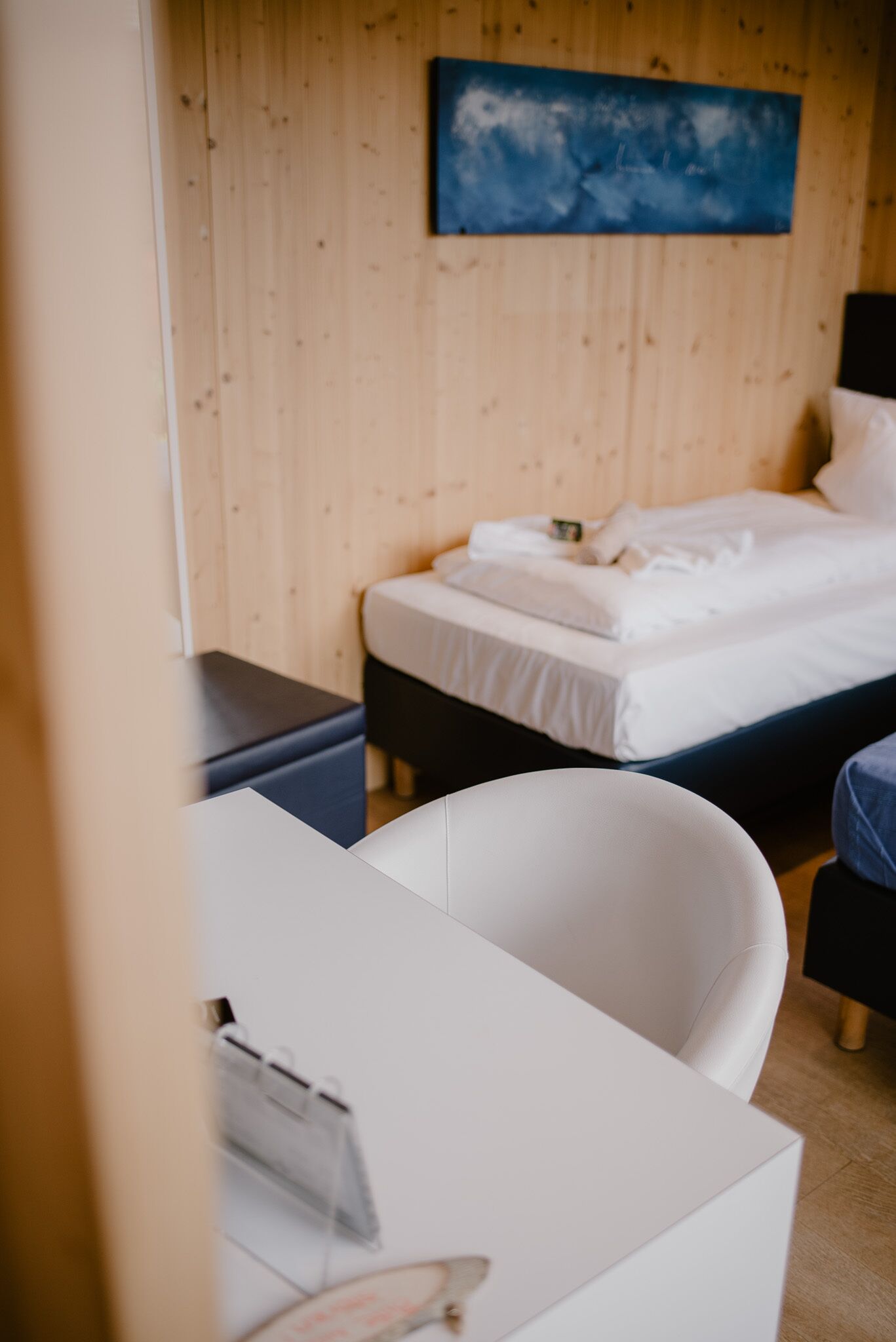 A hotel room with a wooden wall, a bed with white bedding, a desk with a chair, and a blue abstract painting above the bed.