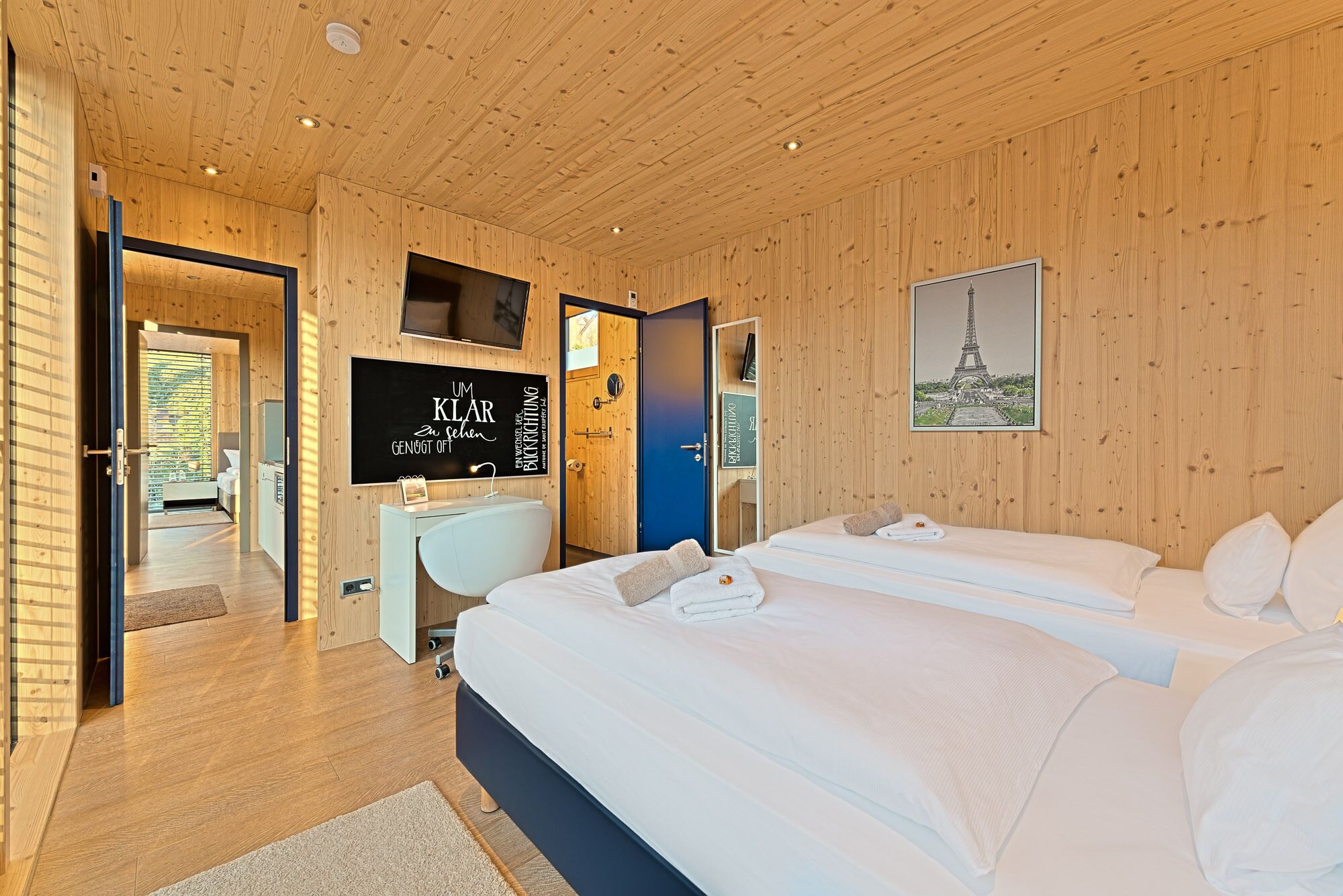 A modern hotel room with wooden paneling, a large bed, a desk, and a television.