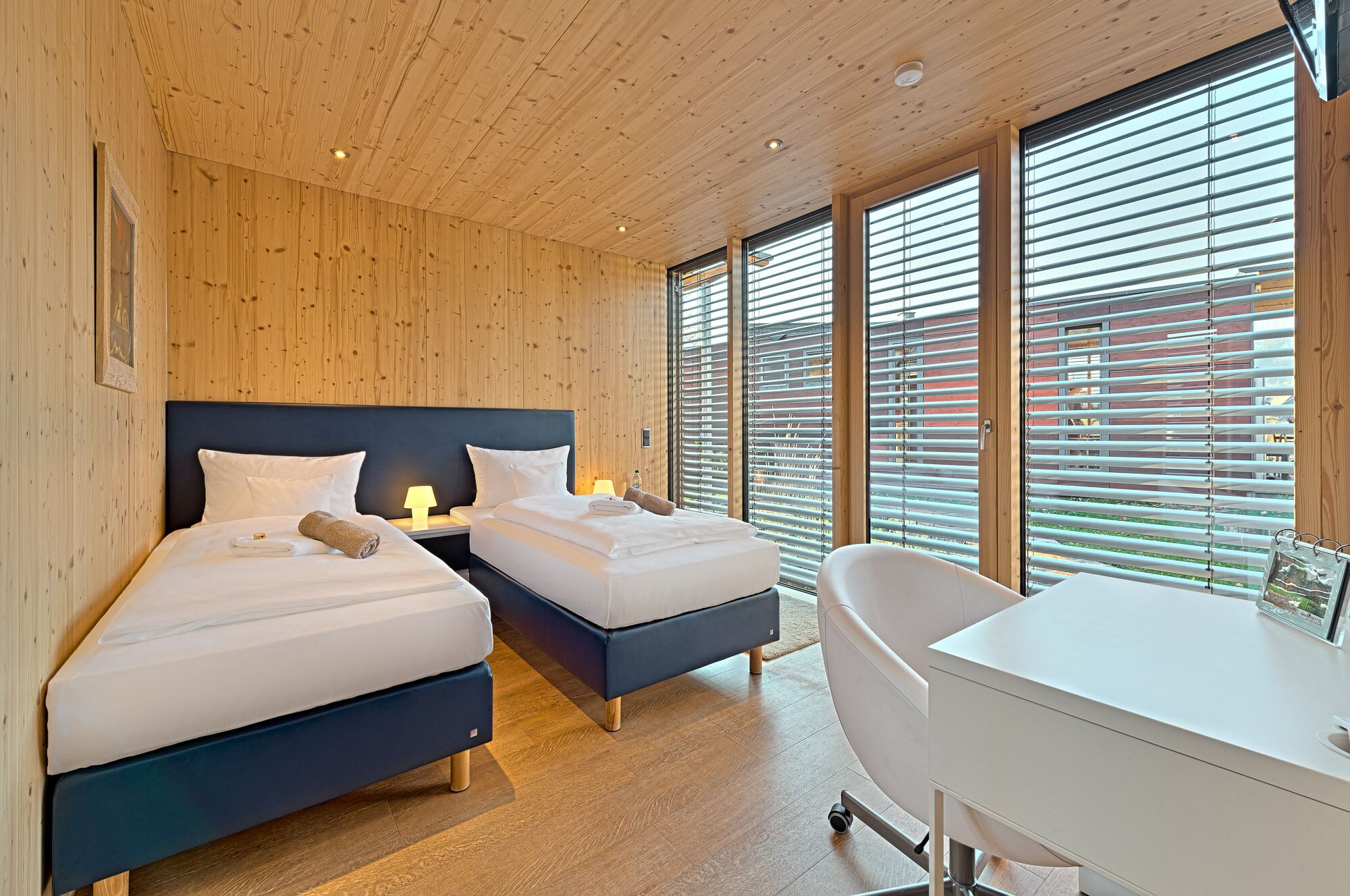 A modern hotel room with two beds, a desk, and a large window with blinds.
