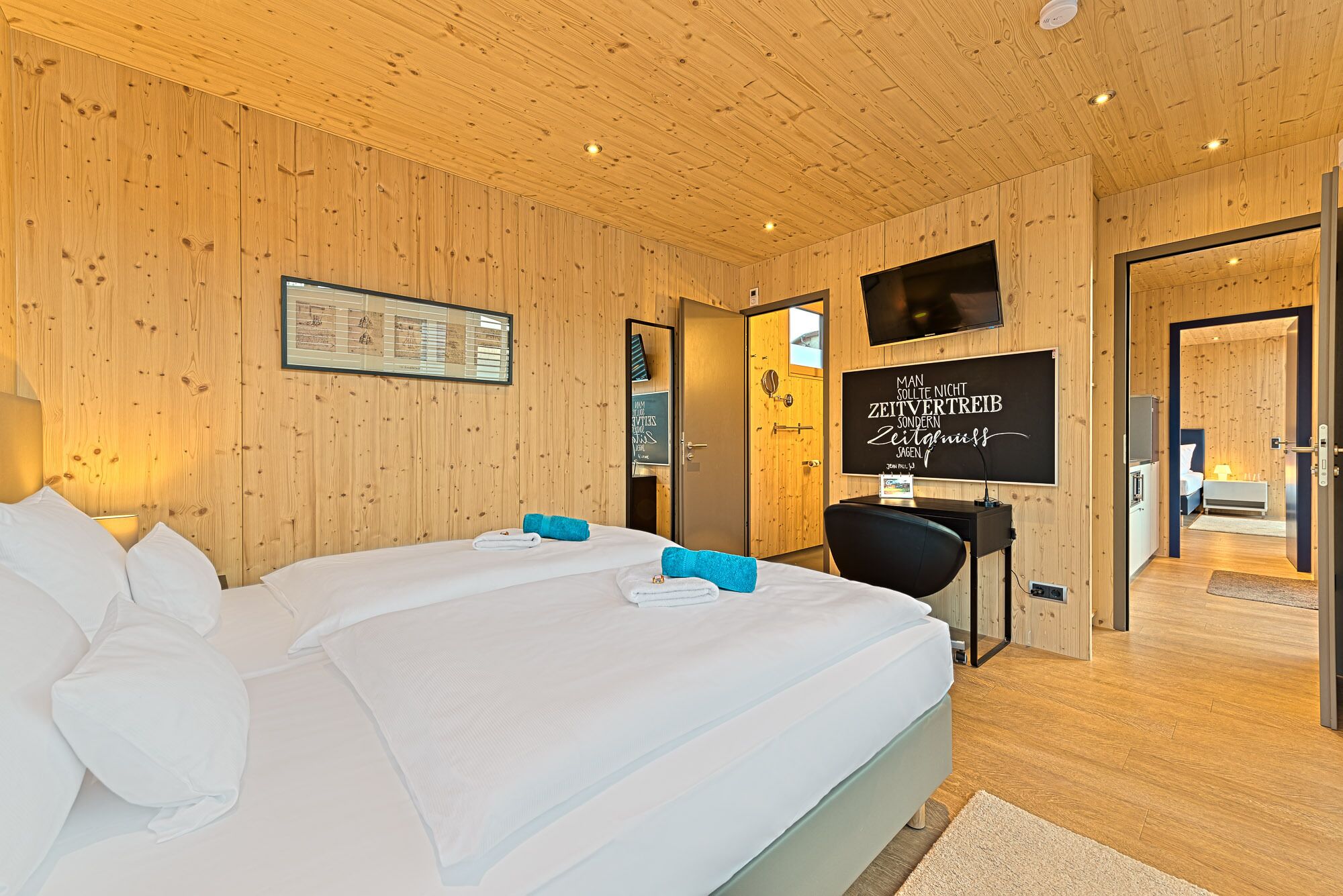 Une chambre d’hôtel avec un plafond en bois, un lit avec du linge de maison blanc, un bureau avec une chaise et un téléviseur fixé au mur.