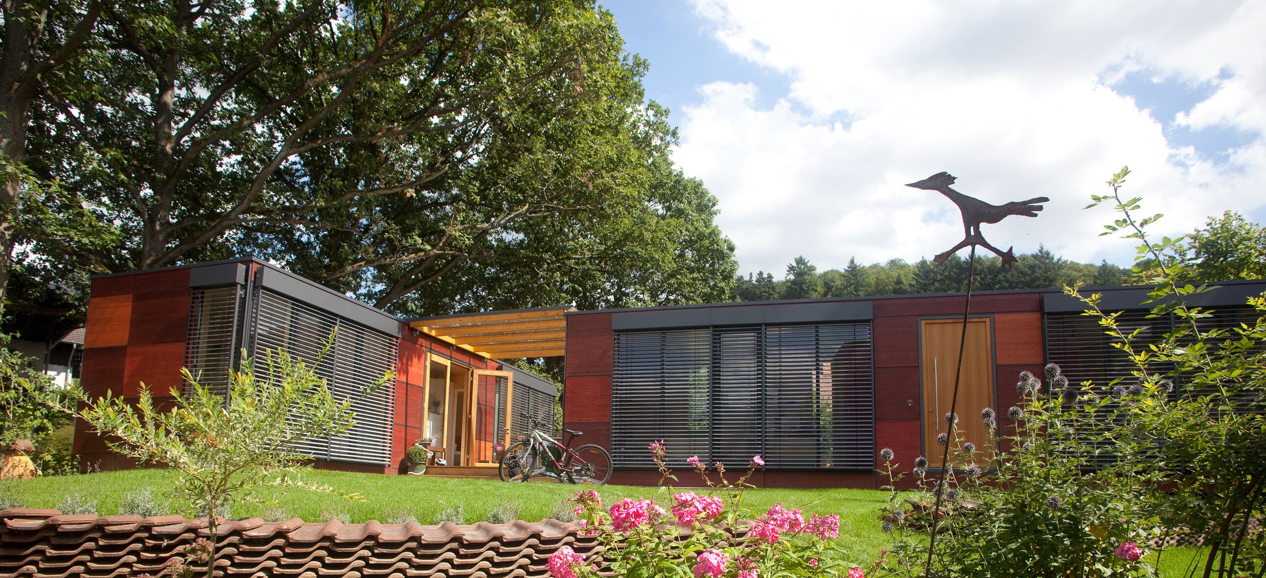 A modern house with a large tree, a bird sculpture, and a garden with flowers.