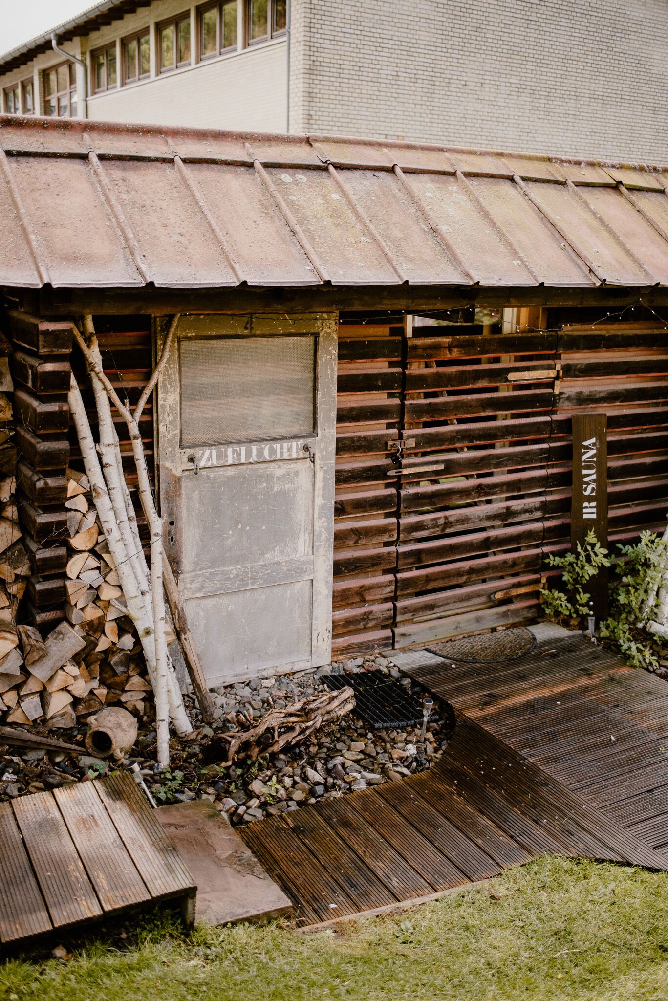 A rustic wooden cabin with a metal roof, a door labeled "SAUNA," and a stack of firewood outside.