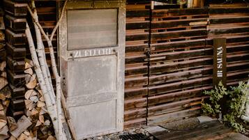 A rustic wooden cabin with a metal roof, a door labeled "SAUNA," and a stack of firewood outside.