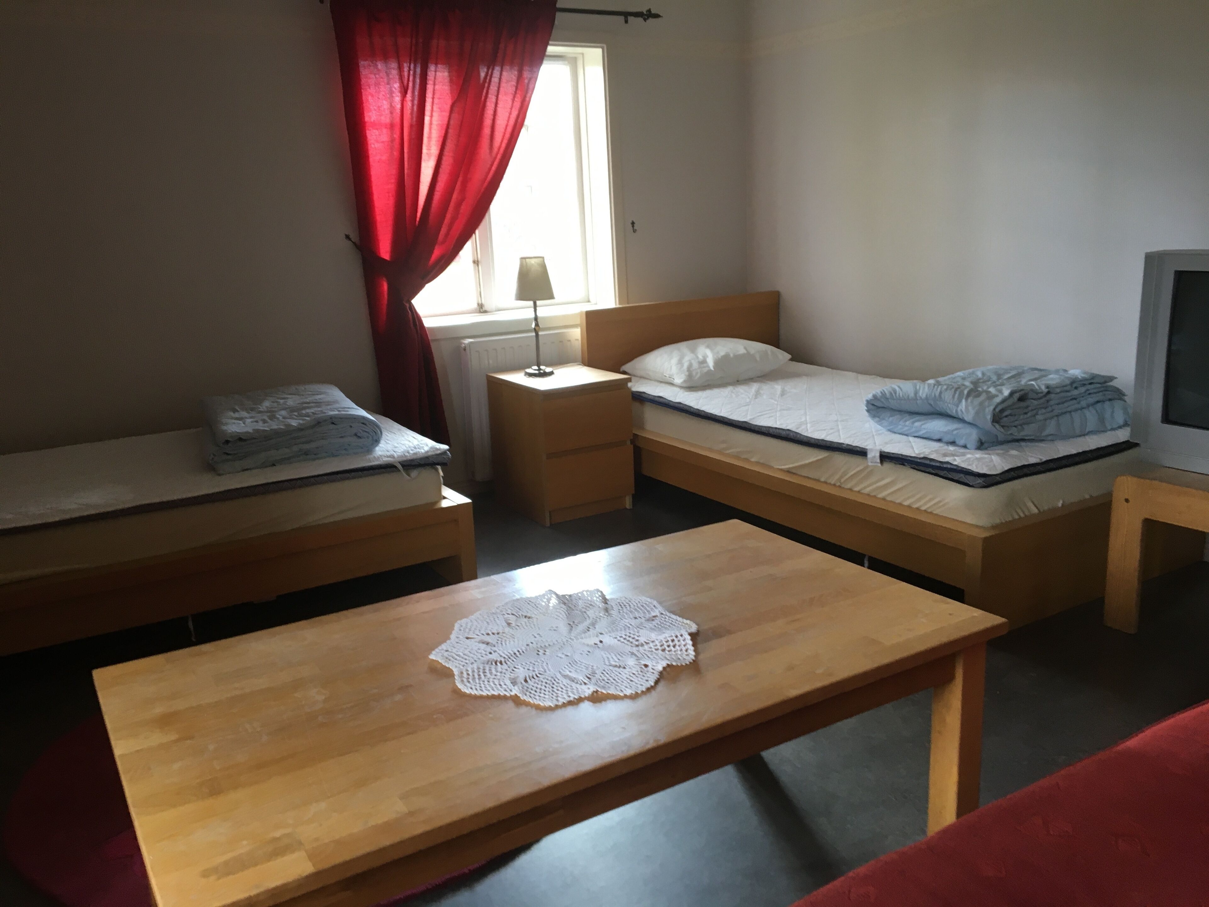 A dormitory room with two single beds, a wooden table with a lace doily, a red curtain, and a window with a lamp.