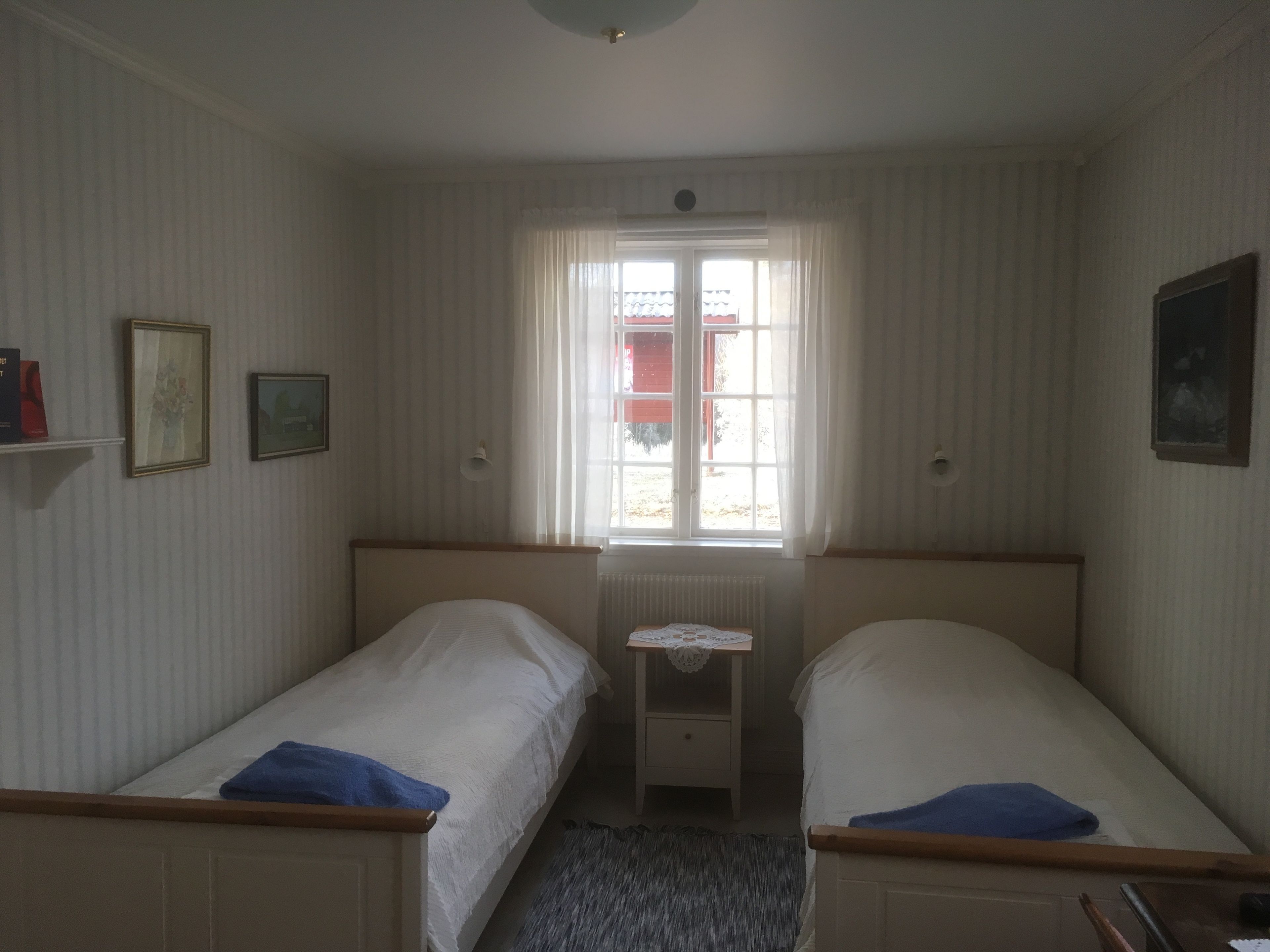 Een kamer met twee eenpersoonsbedden, een klein tafeltje met een kantkleed, en een raam met gordijnen.