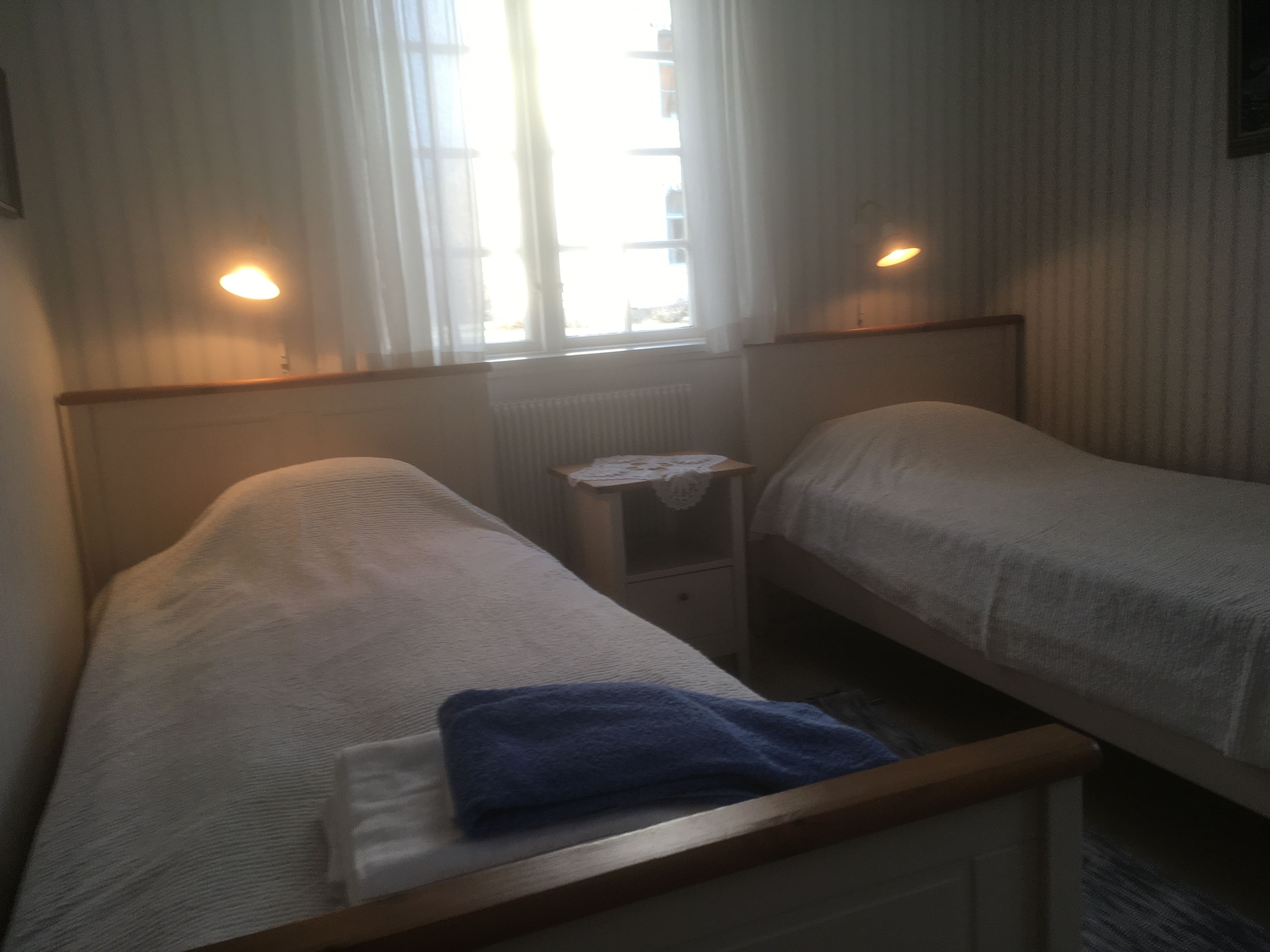 Une petite pièce faiblement éclairée, avec deux lits simples, une petite table et une fenêtre avec des rideaux.