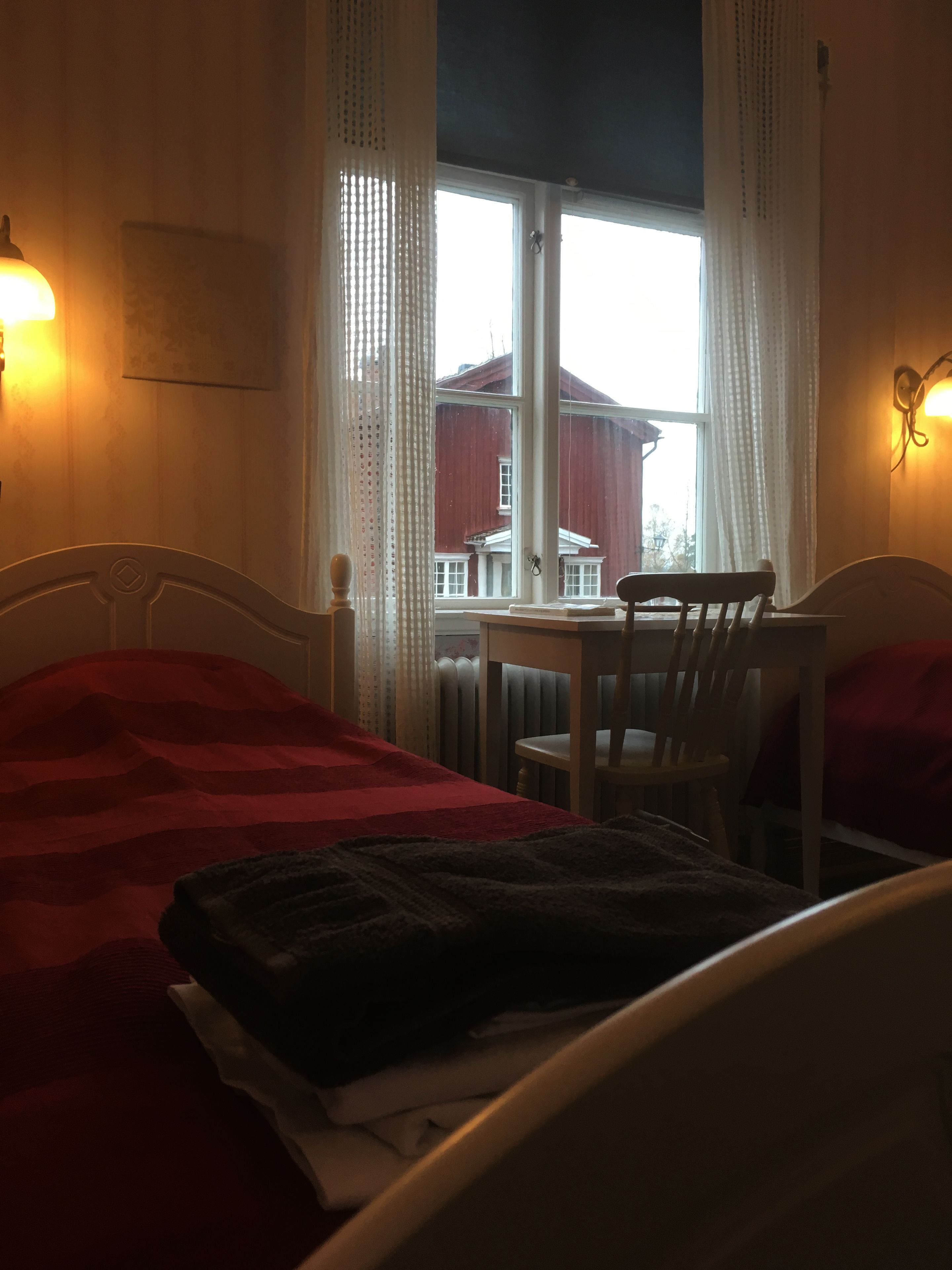 A room with two beds, a table, and chairs. There is a window with curtains, and a lamp on the wall.