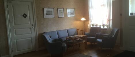 A living room with a sofa, two armchairs, a coffee table, and a lamp. There are three framed pictures on the wall, a door, and a window with curtains.