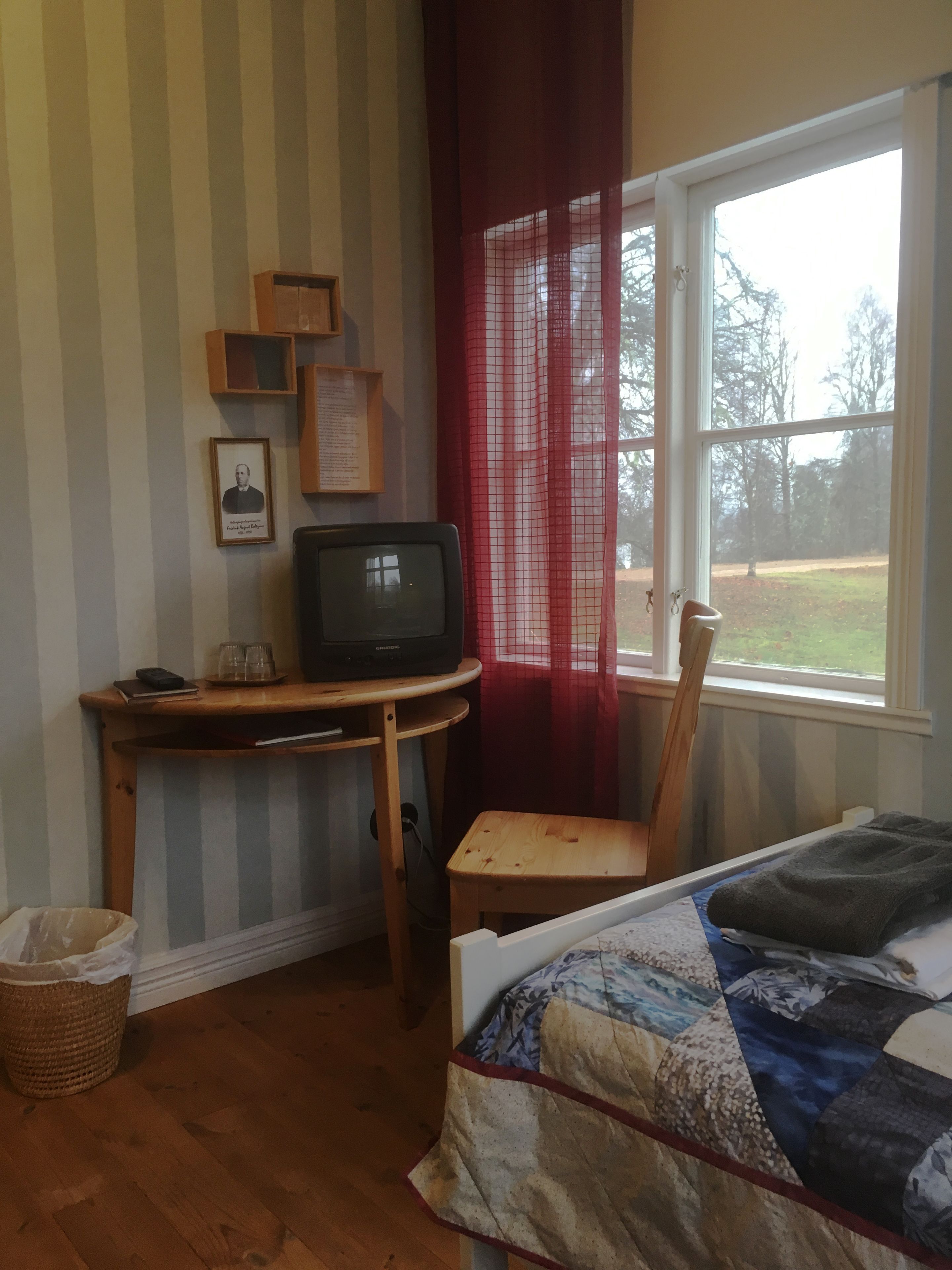 Una habitación con una cama, un escritorio con televisor, una ventana con vistas a árboles y una cortina roja.