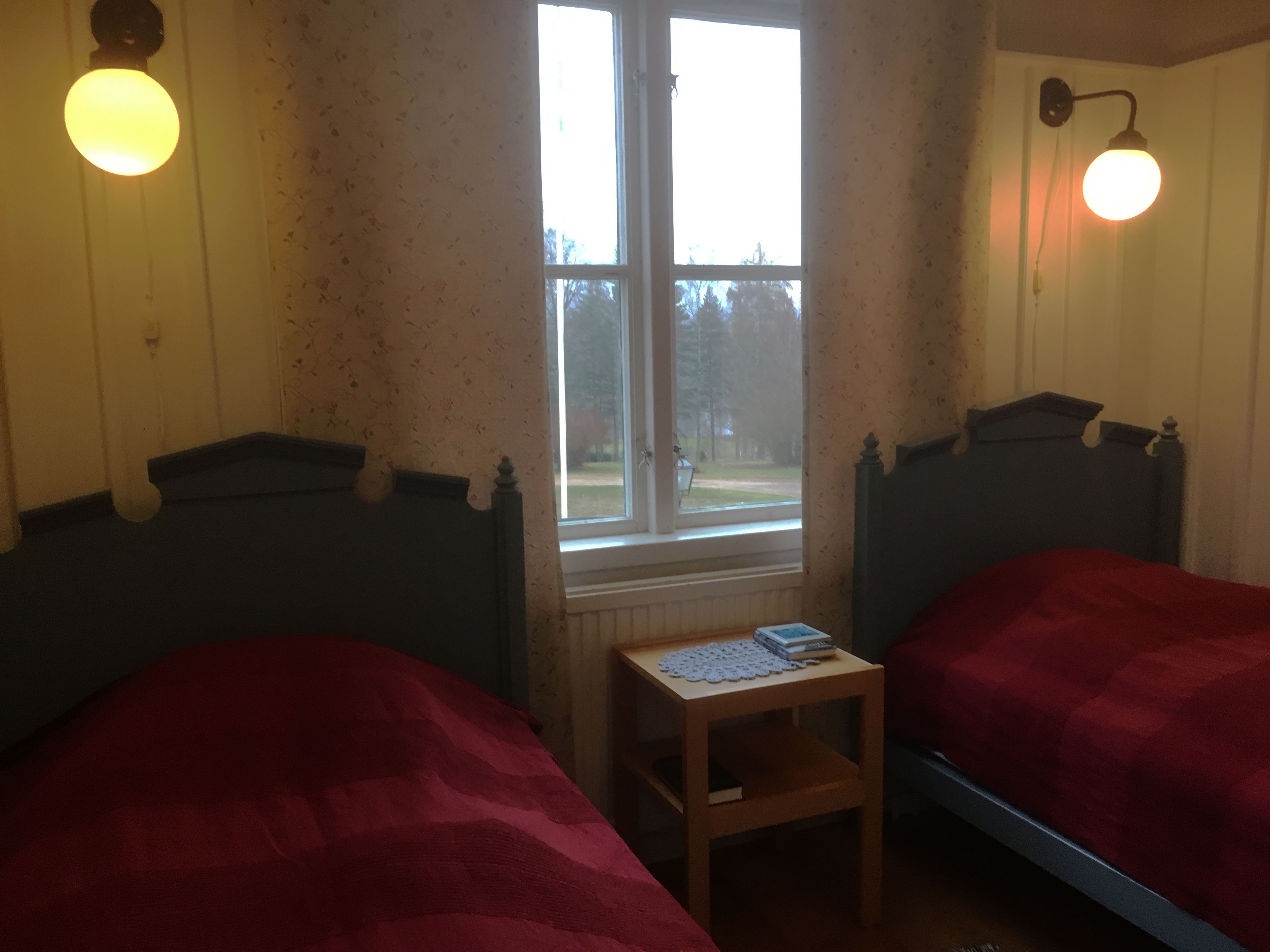 A room with two beds, a small table, a window, and wall-mounted lights.