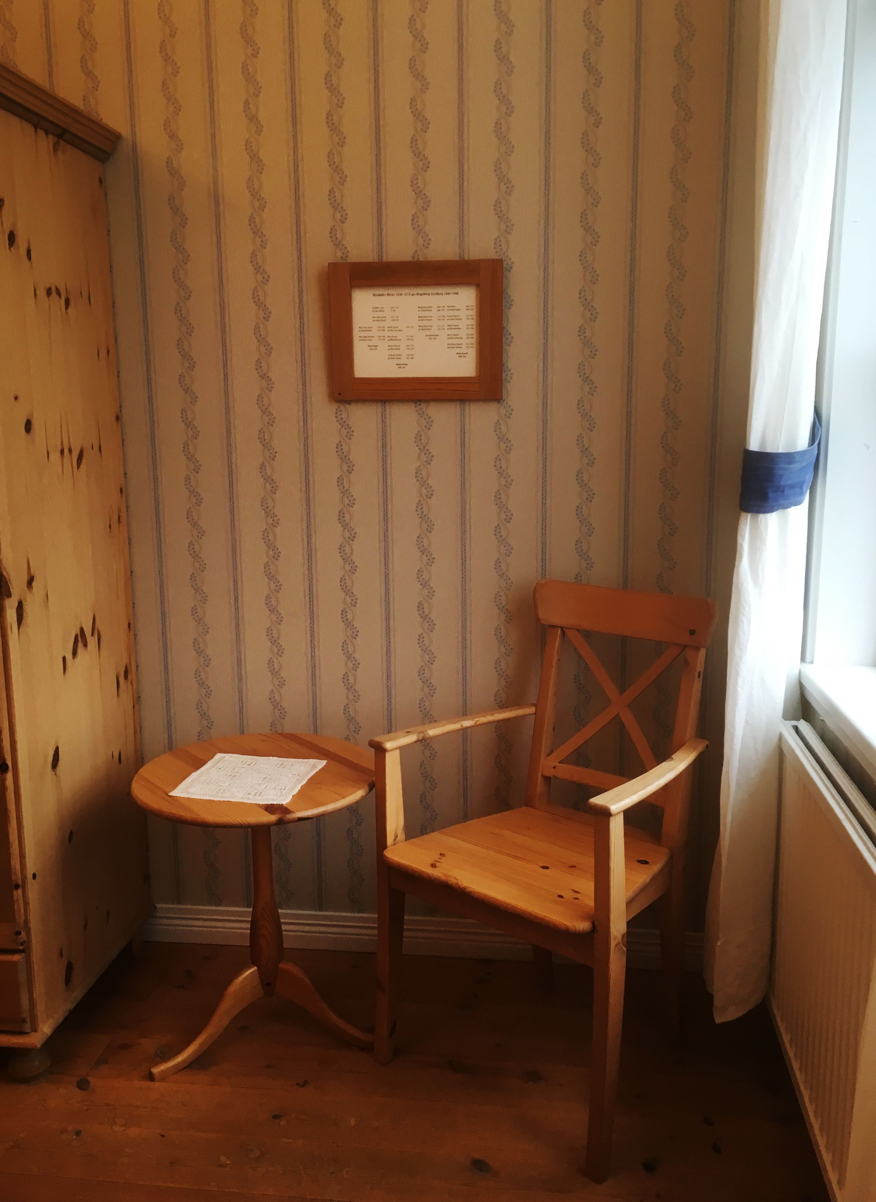 Una habitación de esquina con una silla de madera y una pequeña mesa redonda, un aviso en un marco en la pared, y una ventana con cortinas.