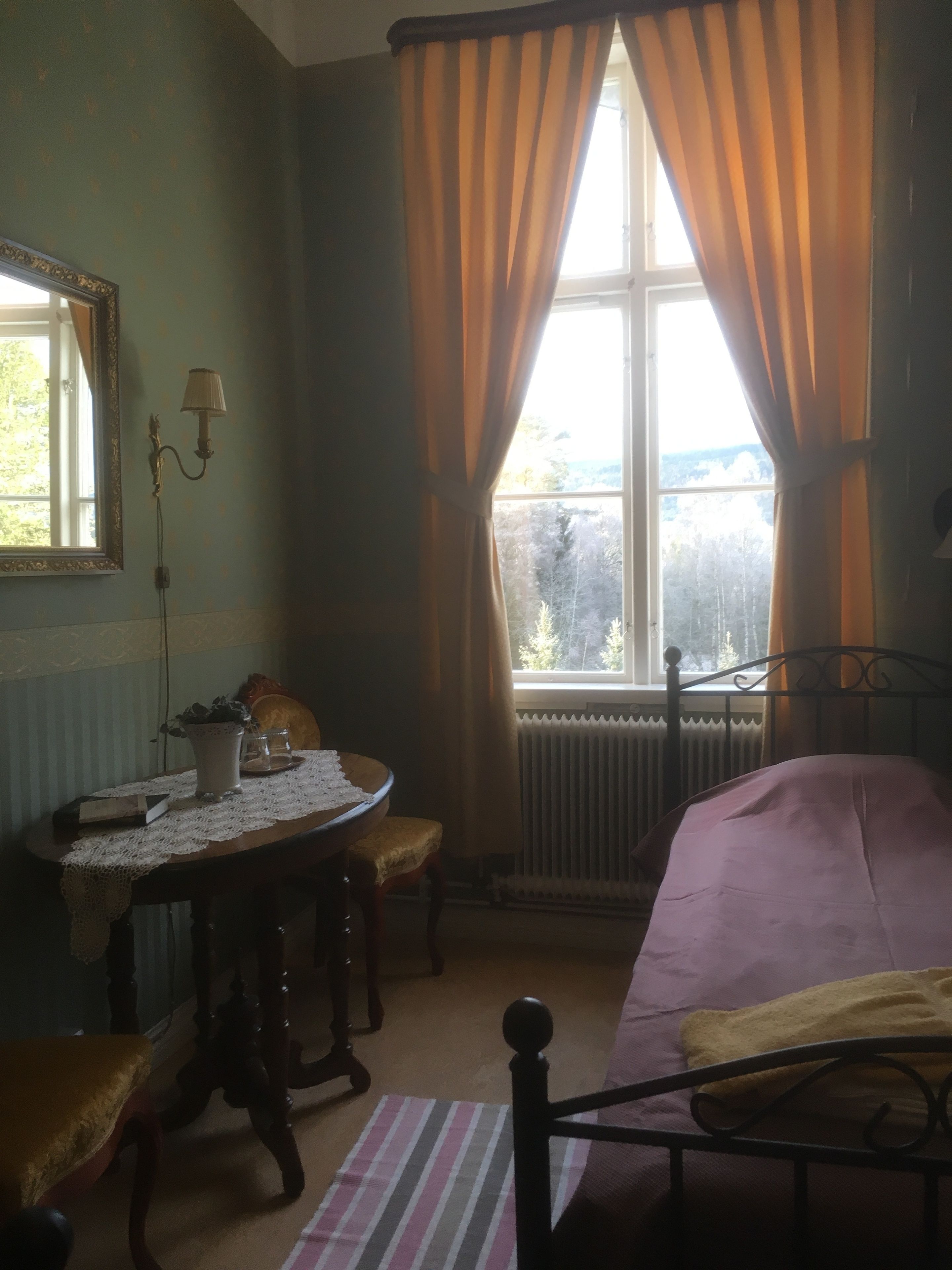 A room with a bed, a table with a lace tablecloth, a chair, a window with curtains, a radiator, and a lamp.