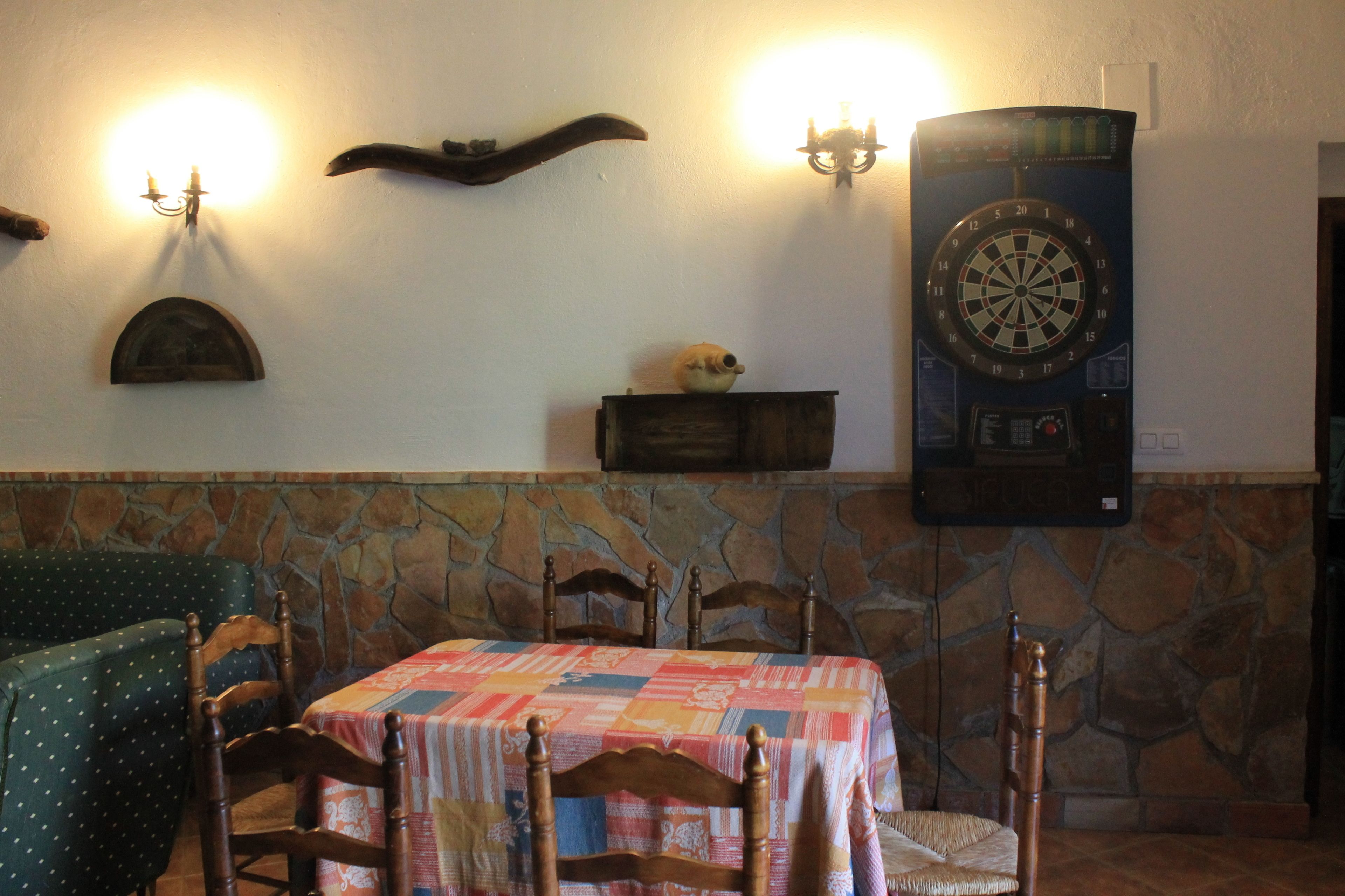 Comedor con una mesa preparada para cuatro personas, pared de piedra, sillas de madera y un dardo en la pared.
