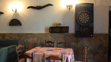 Comedor con una mesa preparada para cuatro personas, pared de piedra, sillas de madera y un dardo en la pared.