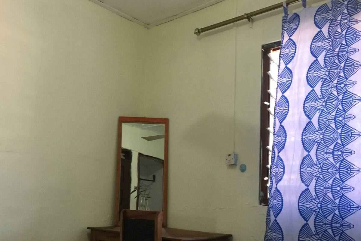 A bedroom with a bed, a desk, a chair, a ceiling fan, and a window with a curtain.