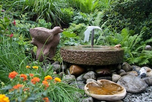 Um jardim com uma fonte de pedra, rodeado por pedras e diversas plantas.