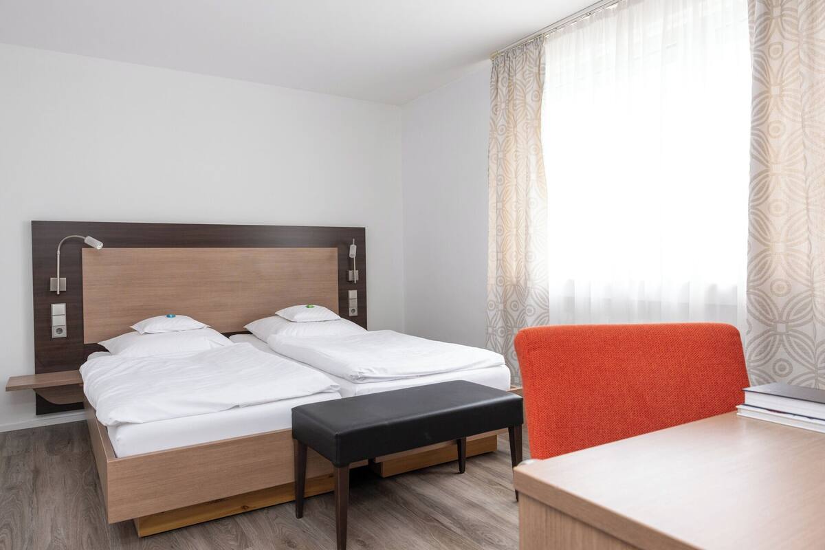 Hotellihuone, jossa on parivuode, yöpöydät, työpöytä tuolilla sekä ikkuna, jossa on verhot.