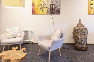 Quarto moderno com duas poltronas brancas, uma mesa de centro de madeira, uma estátua de Buda e quadros abstratos nas paredes.