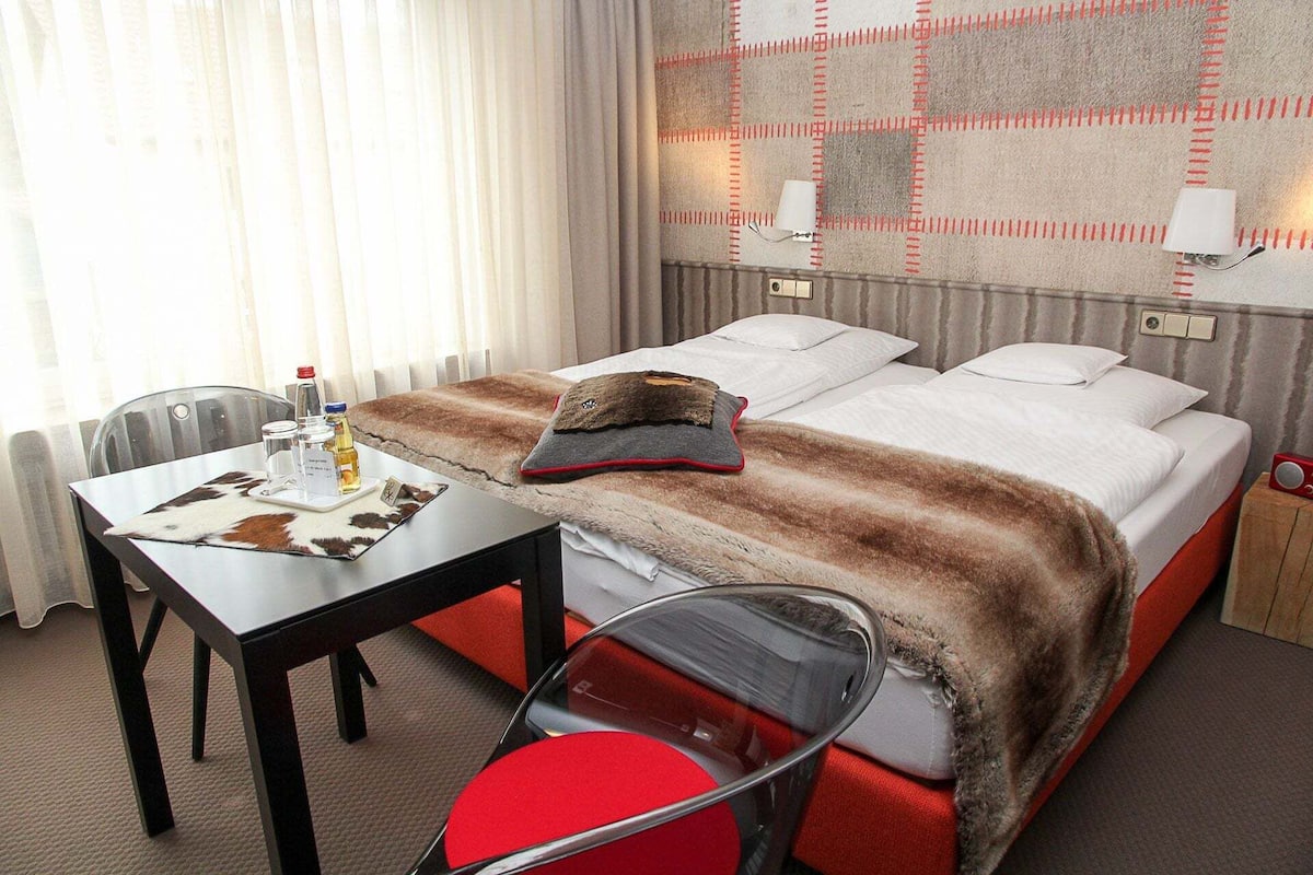 Hotellihuone, jossa on sänky, työpöytä, jonka päällä on pullo, tuoli ja seinässä oleva lamppu.
