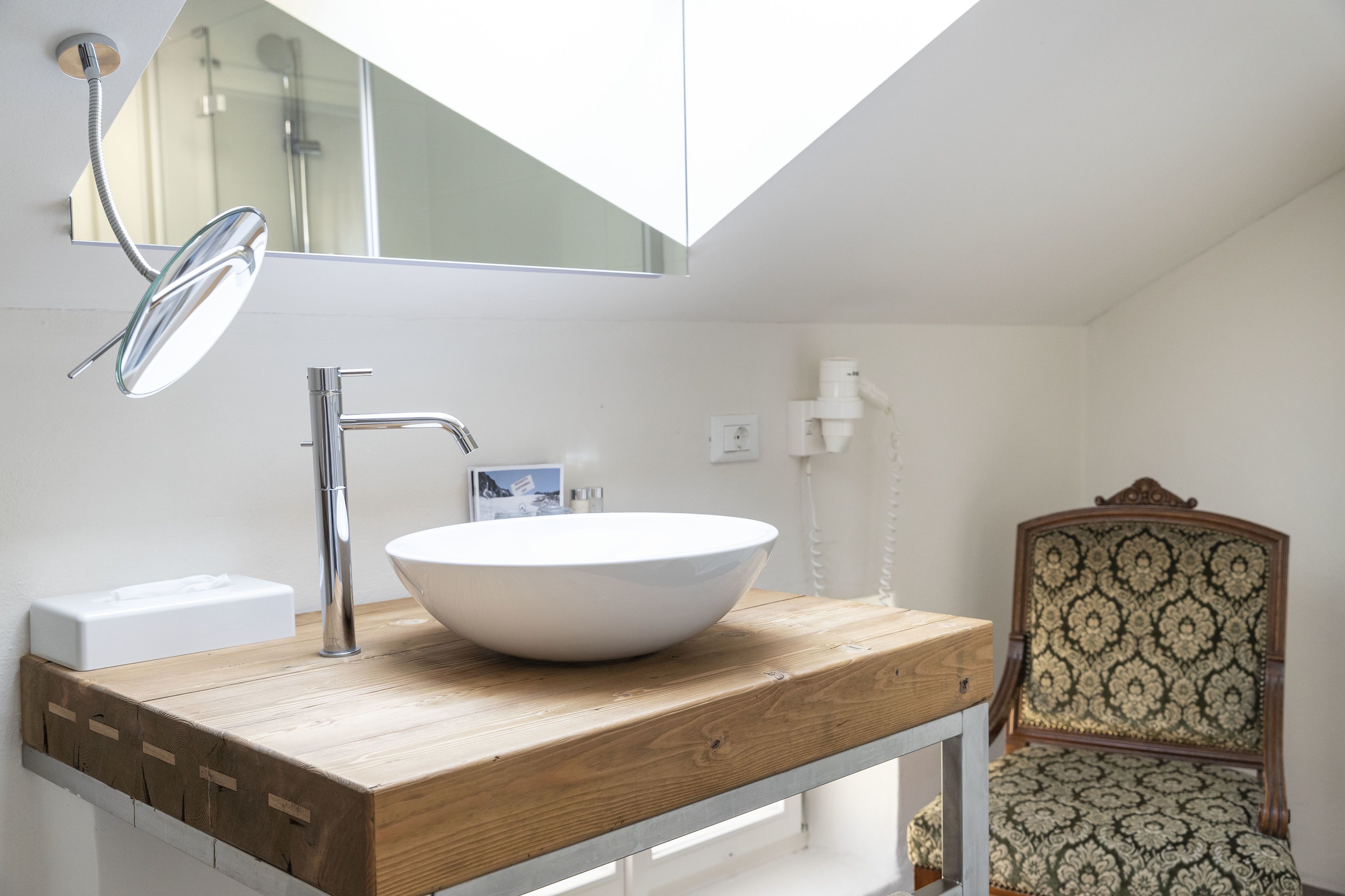 Une salle de bain moderne avec un puits de lumière, un meuble-lavabo en bois, un lavabo blanc et une chaise ancienne.