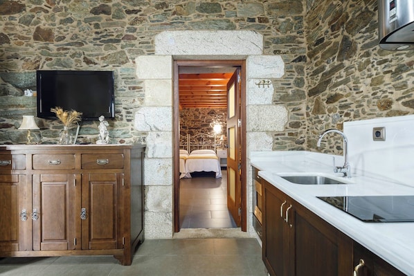 Habitación con paredes de piedra, una cama, un televisor y una zona de cocina con un lavabo y armarios.