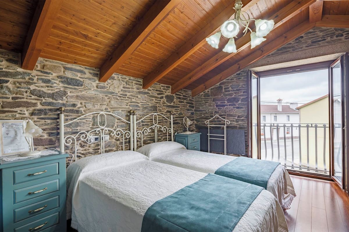 Une chambre avec un mur en pierre, des poutres en bois au plafond, un lit avec une literie blanche et turquoise, une table de chevet et une fenêtre donnant sur des immeubles.