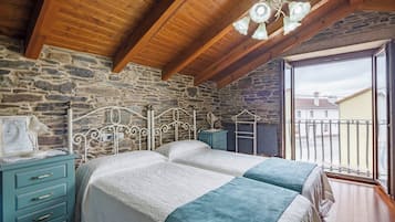 Slaapkamer met een stenen muur, houten balken in het plafond, een bed met wit en teal beddengoed, een nachtkastje en een raam met uitzicht op gebouwen.