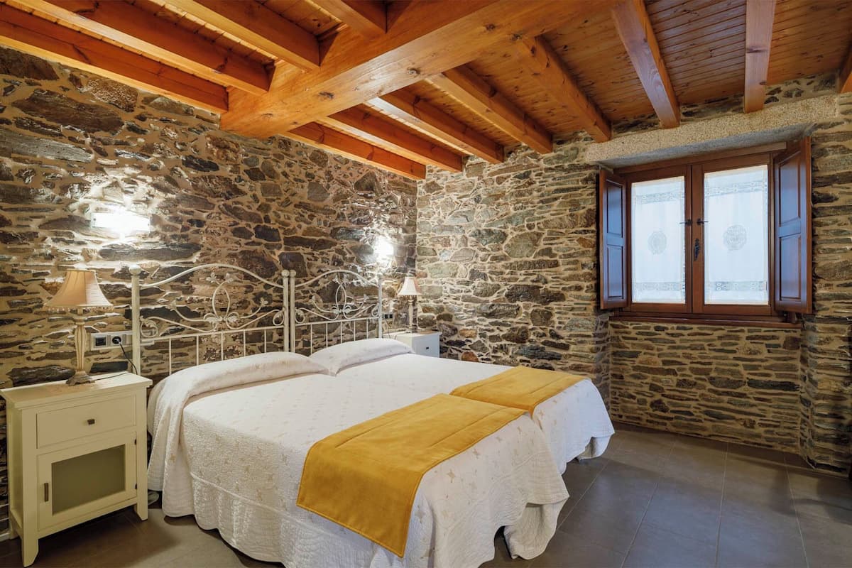 Une chambre avec un mur en pierre, des poutres en bois, un lit recouvert d’une couverture jaune, une table de chevet et une fenêtre avec des rideaux.