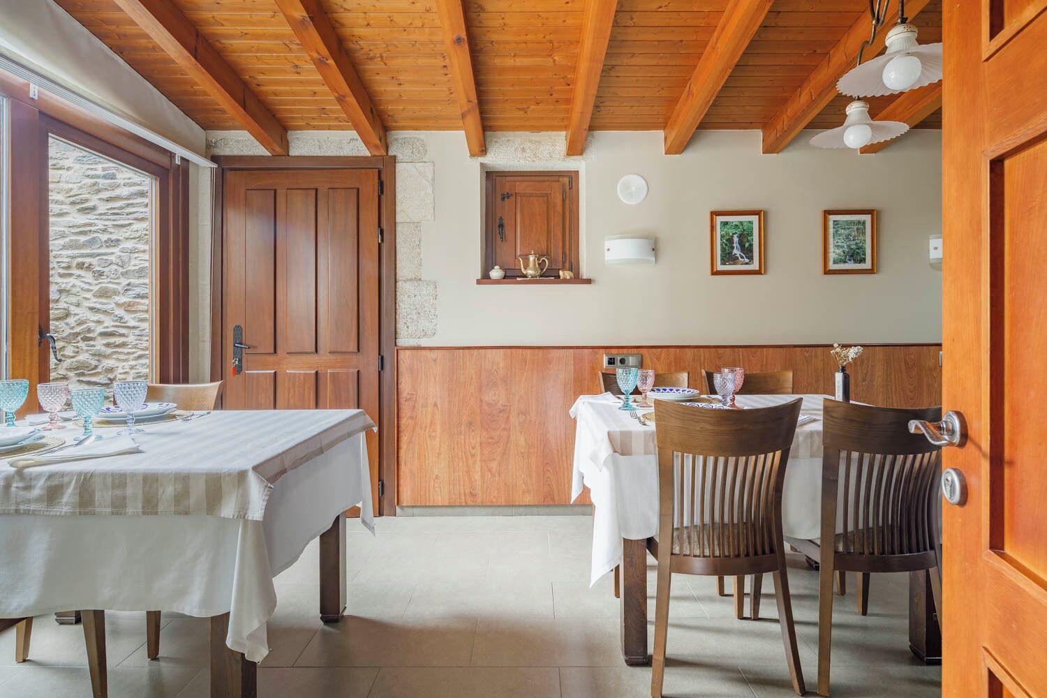 A dining area with wooden tables and chairs, stone walls, and a wooden ceiling.