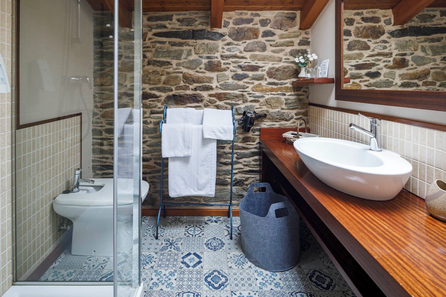 A bathroom with a stone wall, a white sink, a glass shower enclosure, and a patterned floor.