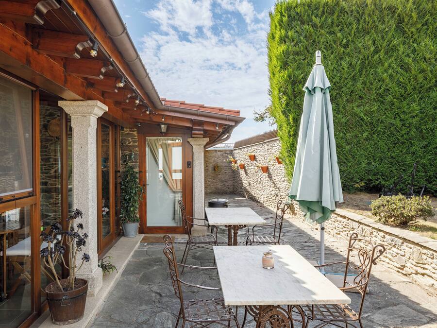 Een terras buiten met stenen vloer, metalen stoelen en tafels. Er staan plantenpotten en een groene parasol.
