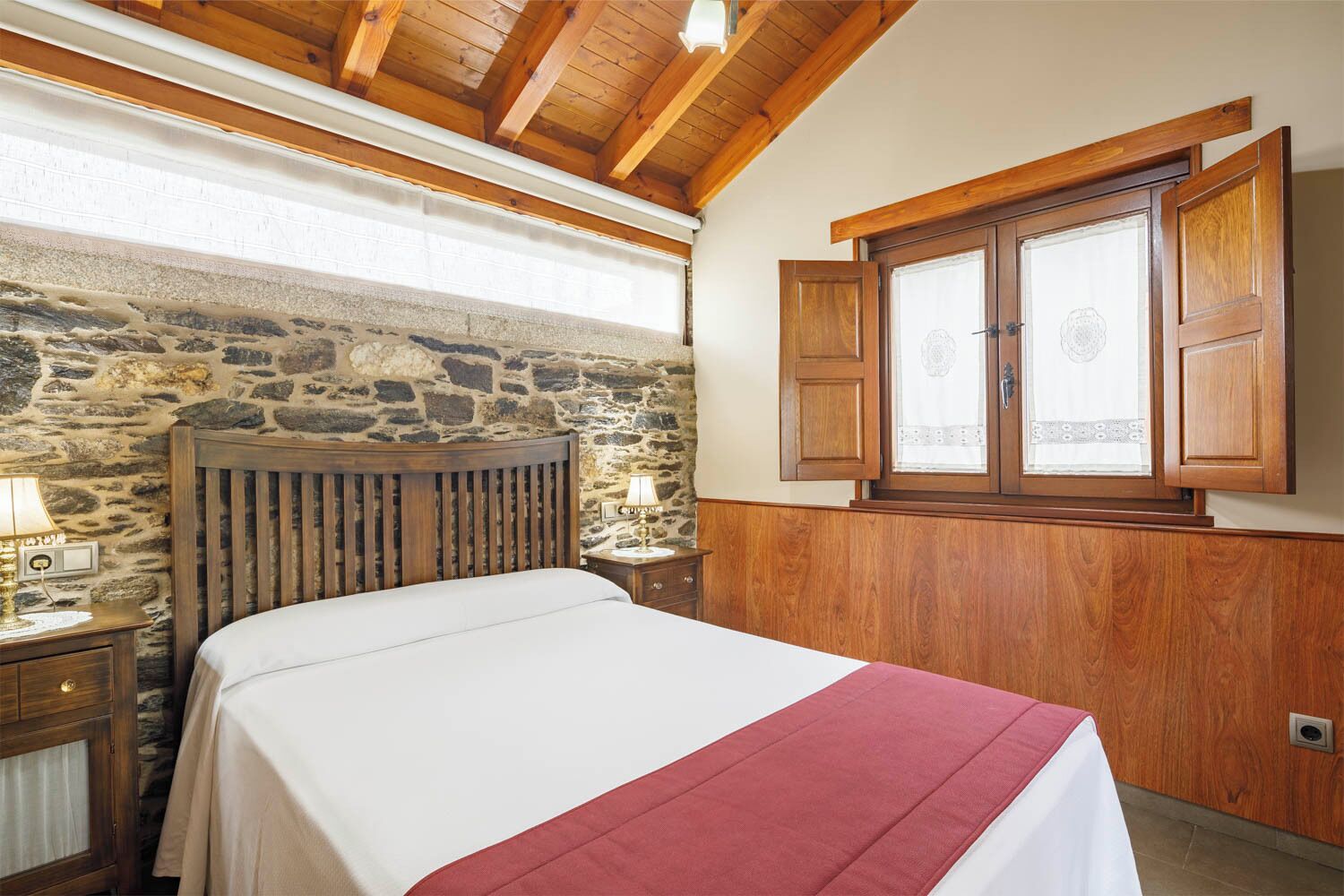 A bedroom with a stone wall, wooden ceiling beams, a bed with a white and red bedspread, a nightstand with a lamp, and a window with wooden shutters.