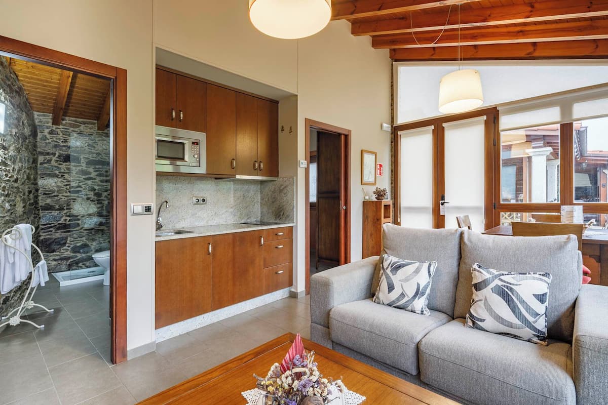 Une cuisine moderne avec des meubles en bois et un mur en pierre, un canapé gris agrémenté de coussins à motifs et une table basse en bois ornée de fleurs.