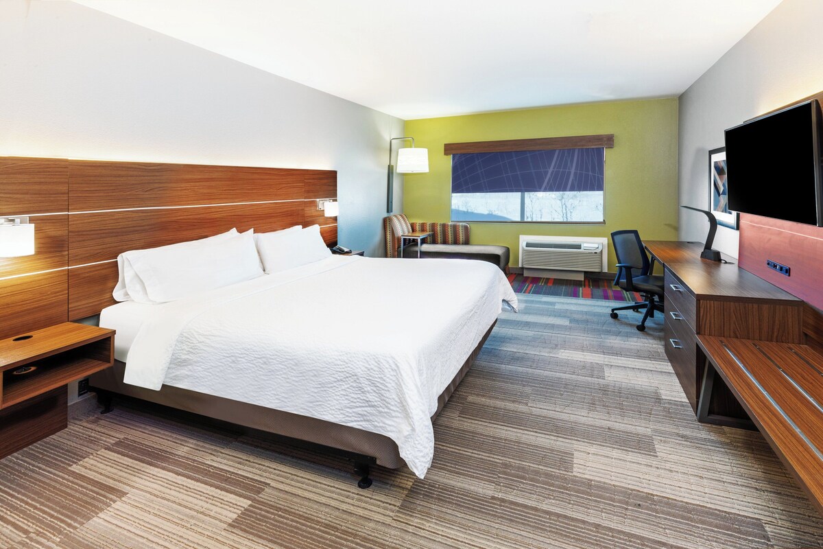 A hotel room with a large bed, a desk with a flat-screen TV, a chair, a window with a view, and a wall-mounted air conditioner.
