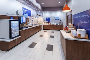 Un espace moderne dédié au petit-déjeuner, équipé d’un buffet, d’un réfrigérateur contenant du lait et d’un comptoir avec des assiettes et des couverts.
