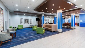 Um hall amplo com carpete azul, sofás verde e bege, uma mesa de centro de madeira e uma televisão presa na parede.