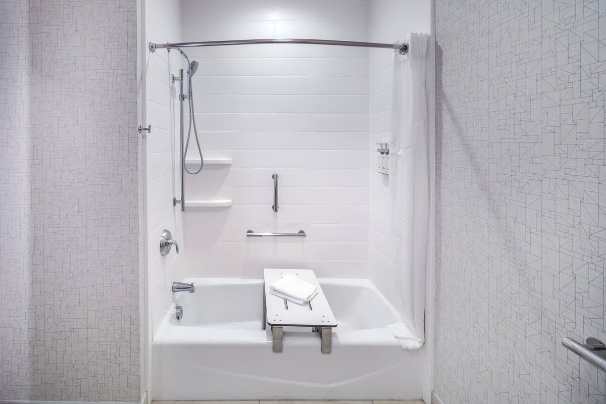 A modern bathroom with a walk-in shower, a bathtub with a small stool, and a handrail.