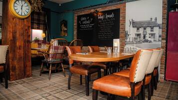 Interior de un restaurante con una mesa y sillas de madera, un reloj en la pared y una pizarra con las especialidades.