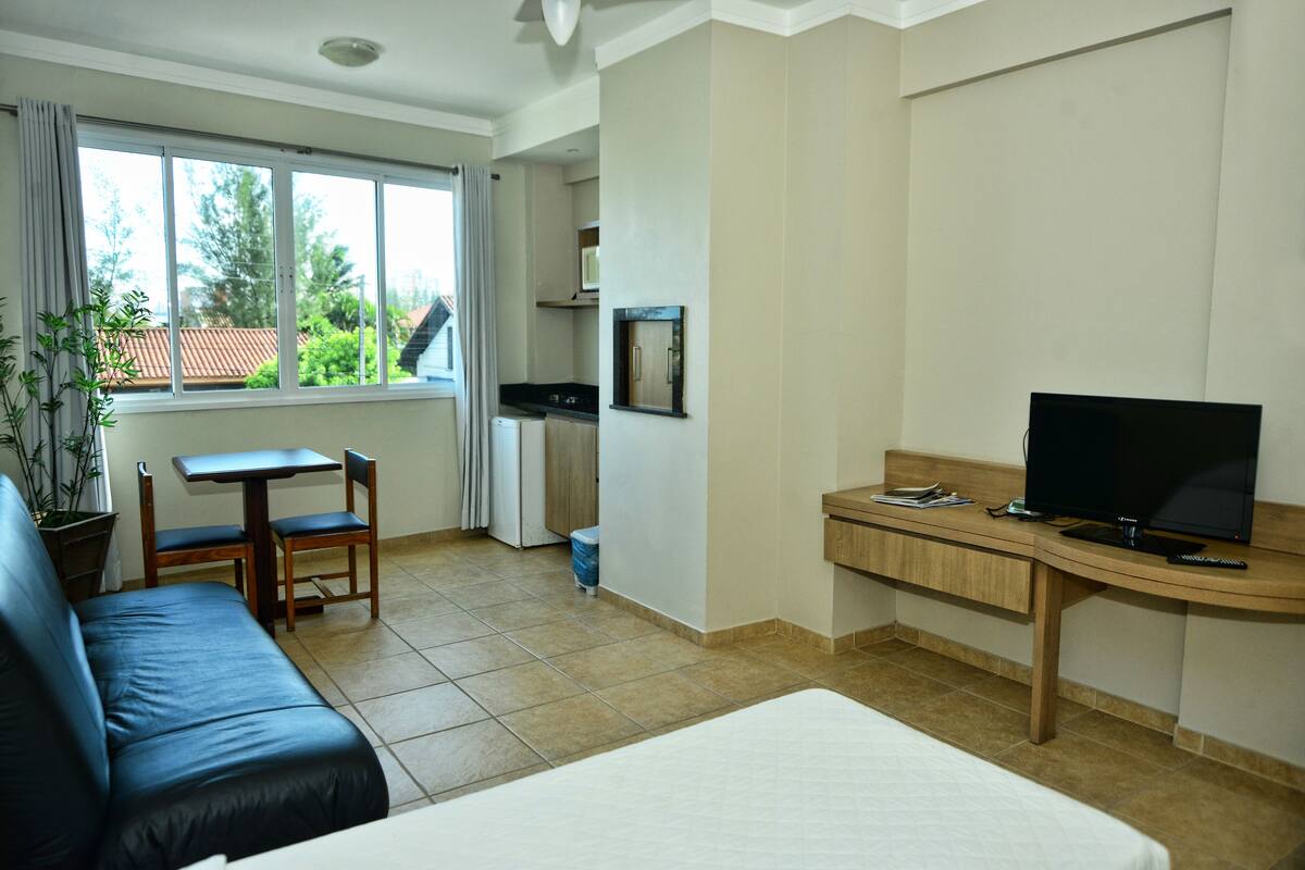 A hotel room with a bed, a desk with a TV, a small kitchen area, and a window with curtains.