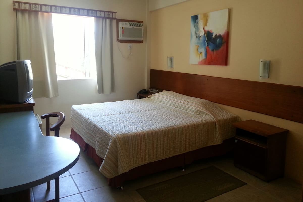 A hotel room with a bed, a desk with a television, a window with curtains, an air conditioning unit, and a painting on the wall.