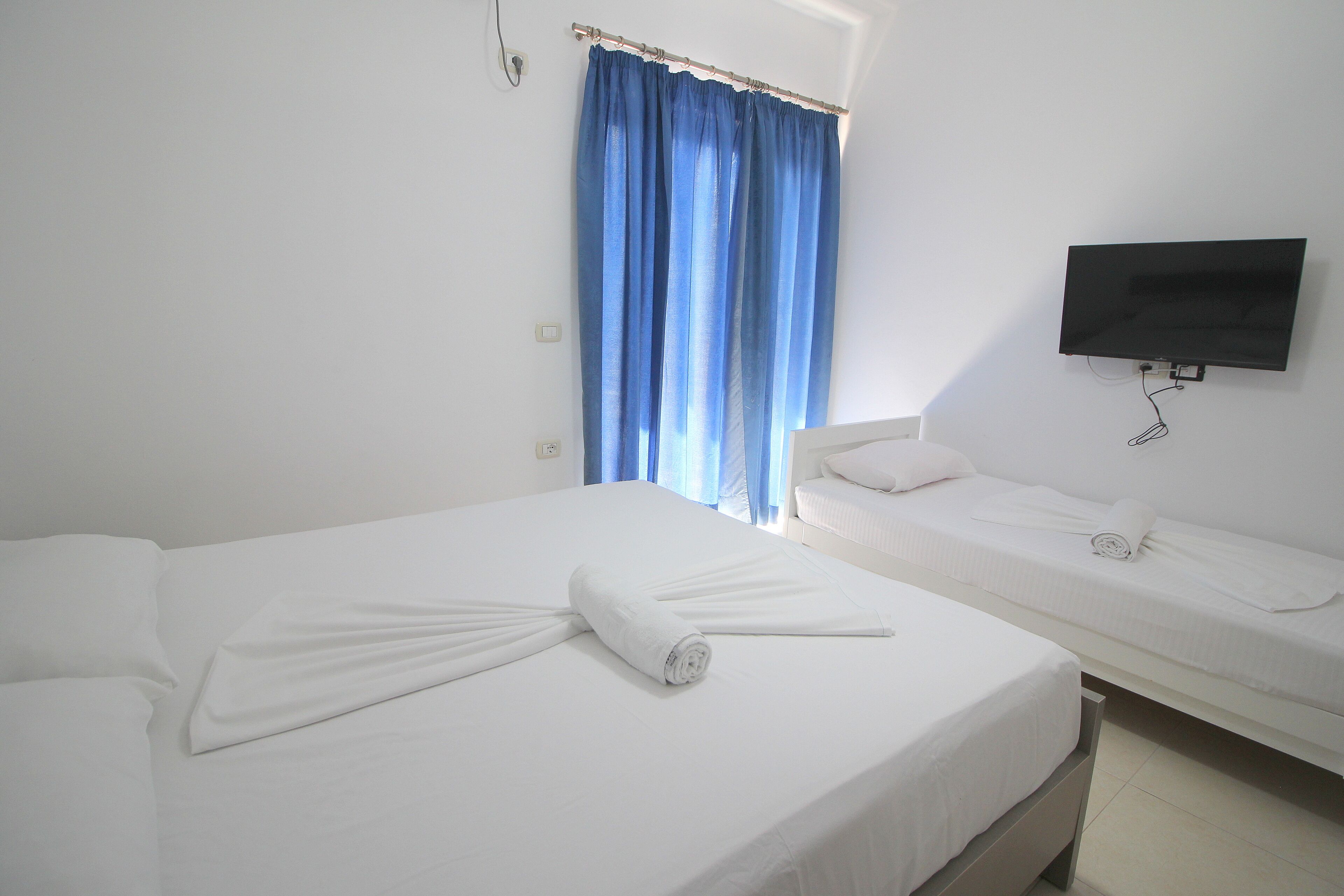 A hotel room with two beds, a TV mounted on the wall, and a window with blue curtains.