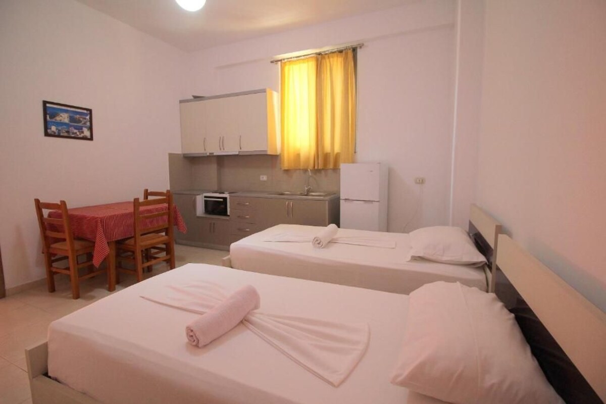 A hotel room with two beds, a small kitchenette, a dining table with chairs, and a window with yellow curtains.