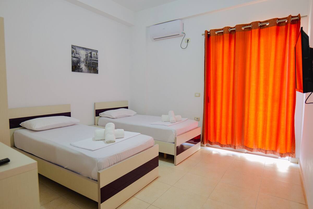 A hotel room with two beds, a small desk, a wall-mounted air conditioner, and a window with orange curtains.