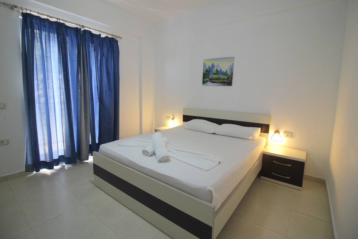 A neatly made bed with white linens and towels, a painting on the wall, and a window with blue curtains.