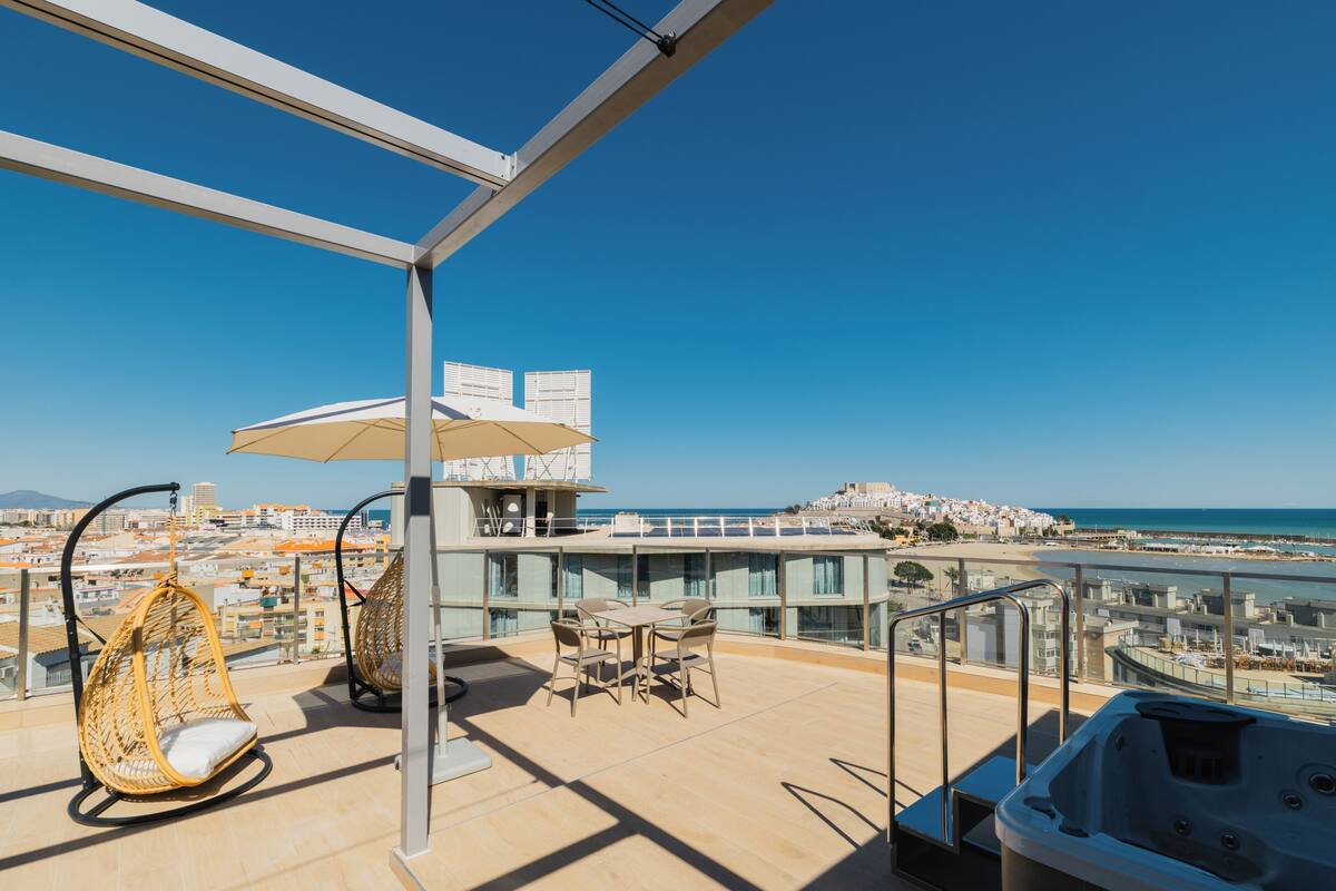 Un balcón con una silla colgante, una mesa con sillas, un jacuzzi y vistas al mar y al paisaje urbano.