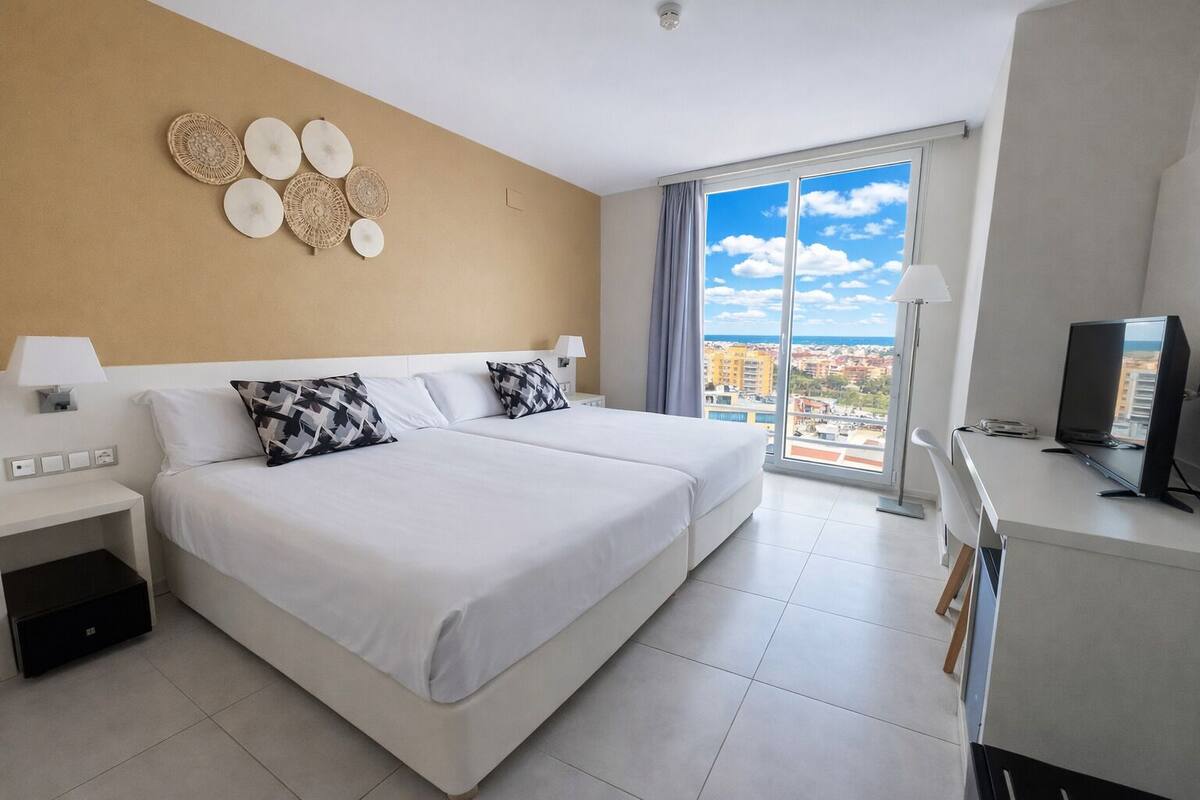 Una habitación de hotel con una cama, un escritorio, un televisor y un amplio ventanal con vista a la ciudad.