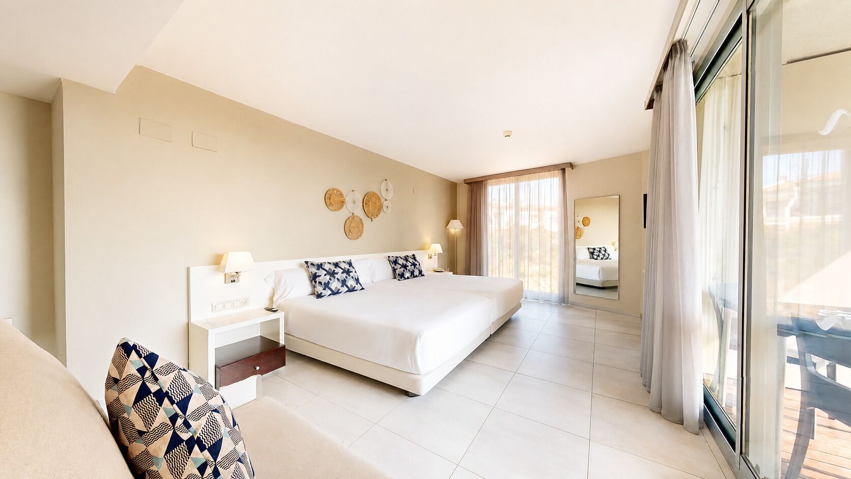 A hotel bedroom with a large bed, bedside tables, a nightstand, a sofa, and a sliding glass door to the balcony.